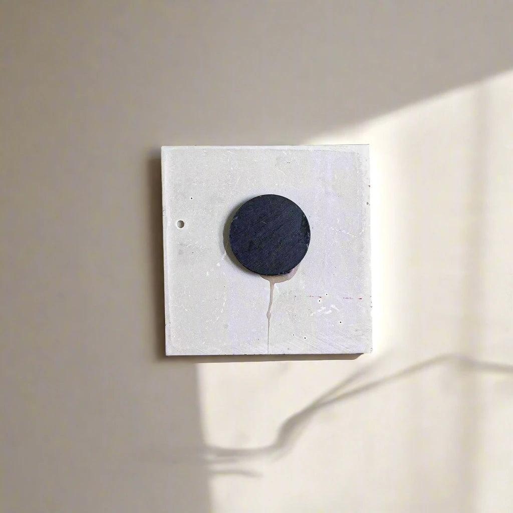 A minimalist artwork featuring a square white canvas with a small, circular, black object resembling a magnet positioned slightly above center. A faint shadow cast by the black circle extends toward the lower right corner, adding depth to the monochromatic composition.