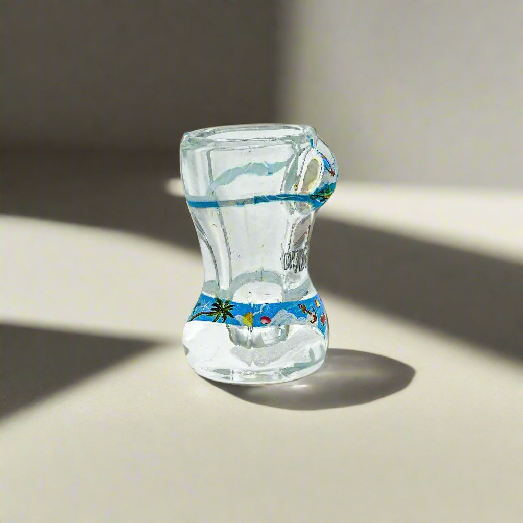 A clear shot glass designed with a sculpted torso in a bikini, showcasing beach and ocean imagery. The bikini is illustrated with scenes of the seaside. The front of the glass displays "I ♥ Algarve" in bold black and red letters, making it an ideal keepsake from the Algarve region.