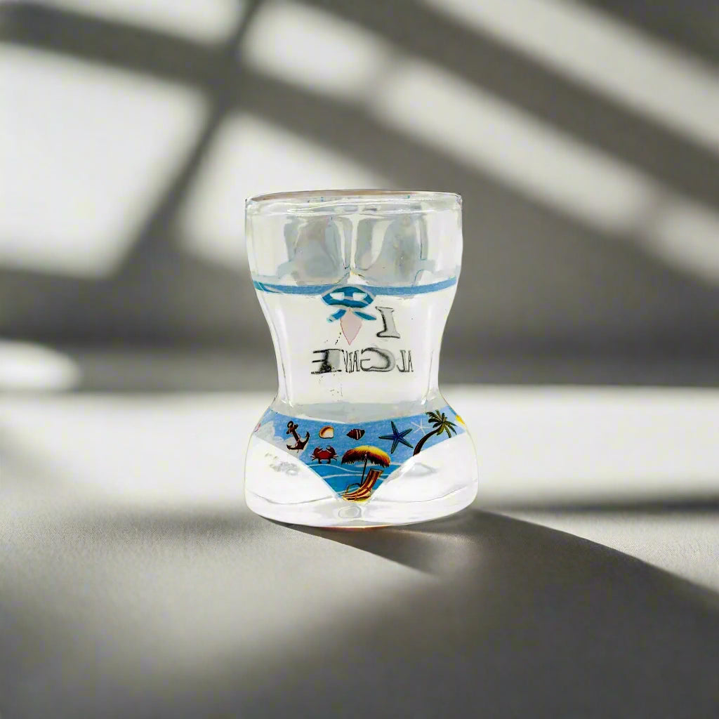A see-through shot glass featuring a torso shape with a bikini illustration, adorned with beach and ocean scenes. The glass is marked with "I ♥ Algarve" in black and red text on the front, serving as a perfect memento from the Algarve area.