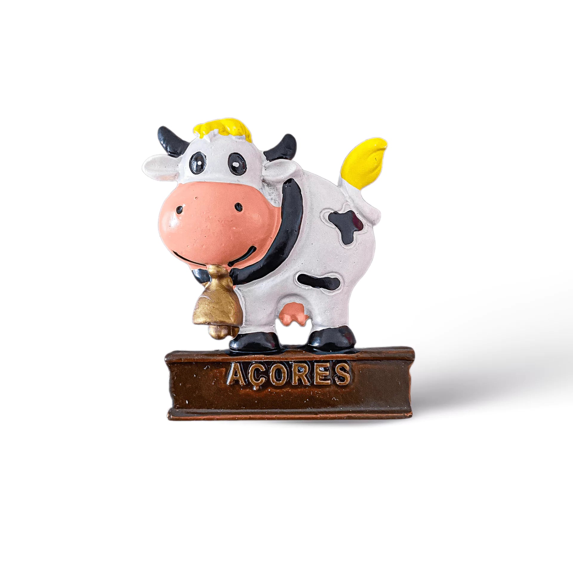 A cheerful cow figurine magnet stands on a brown base with the word "Açores" written on it. The cow is white with black spots, has a yellow tuft of hair and tail, and is holding a small bell in its mouth. This charming souvenir from Portugal features a pure white background.