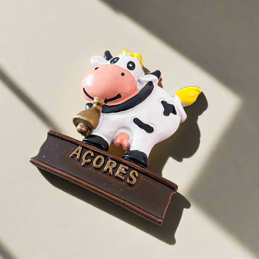 A playful cow figurine magnet sits on a brown base with "Açores" inscribed on it. The cow is white with black spots, a yellow tuft of hair and tail, and is holding a tiny bell in its mouth. This delightful Portugal souvenir stands against a clean white background.