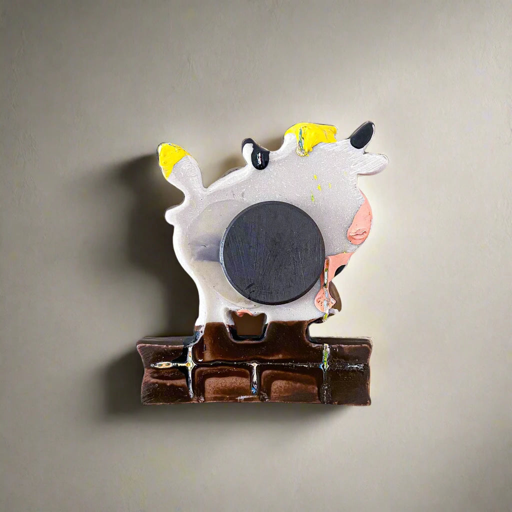 A whimsical cow figurine magnet rests on a brown base featuring the word "Açores." The cow, white with black spots, has a yellow tuft of hair and tail, and holds a small bell in its mouth. This charming souvenir from Portugal is set against a crisp white background.
