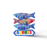 Ceramic artwork featuring three colorful fish with intricate patterns, styled in blue, red, and yellow. Below the fish, a sign reads "AÇORES" in vibrant letters. This charming Portugal souvenir magnet stands out against a plain white background.