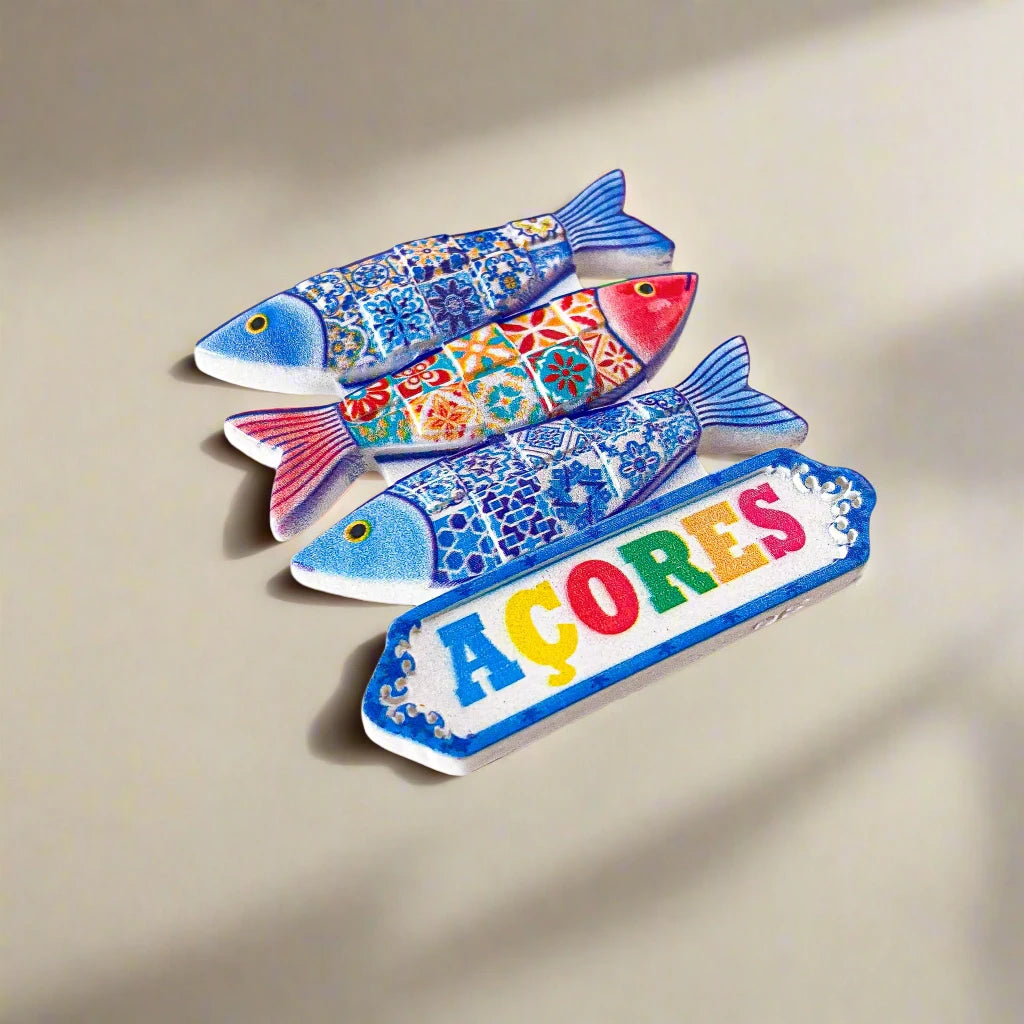 A ceramic artwork displaying three vibrantly colored fish with detailed patterns in blue, red, and yellow. Beneath the fish, a sign reads "AÇORES" in bold, colorful letters. This eye-catching Portugal souvenir magnet is set against a simple white background.