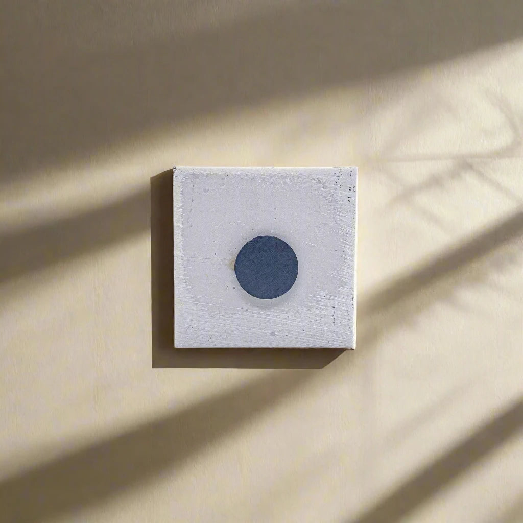 A minimalist artwork featuring a square white canvas with a small, circular, black object resembling a magnet positioned slightly above center. A faint shadow cast by the black circle extends toward the lower right corner, adding depth to the monochromatic composition.