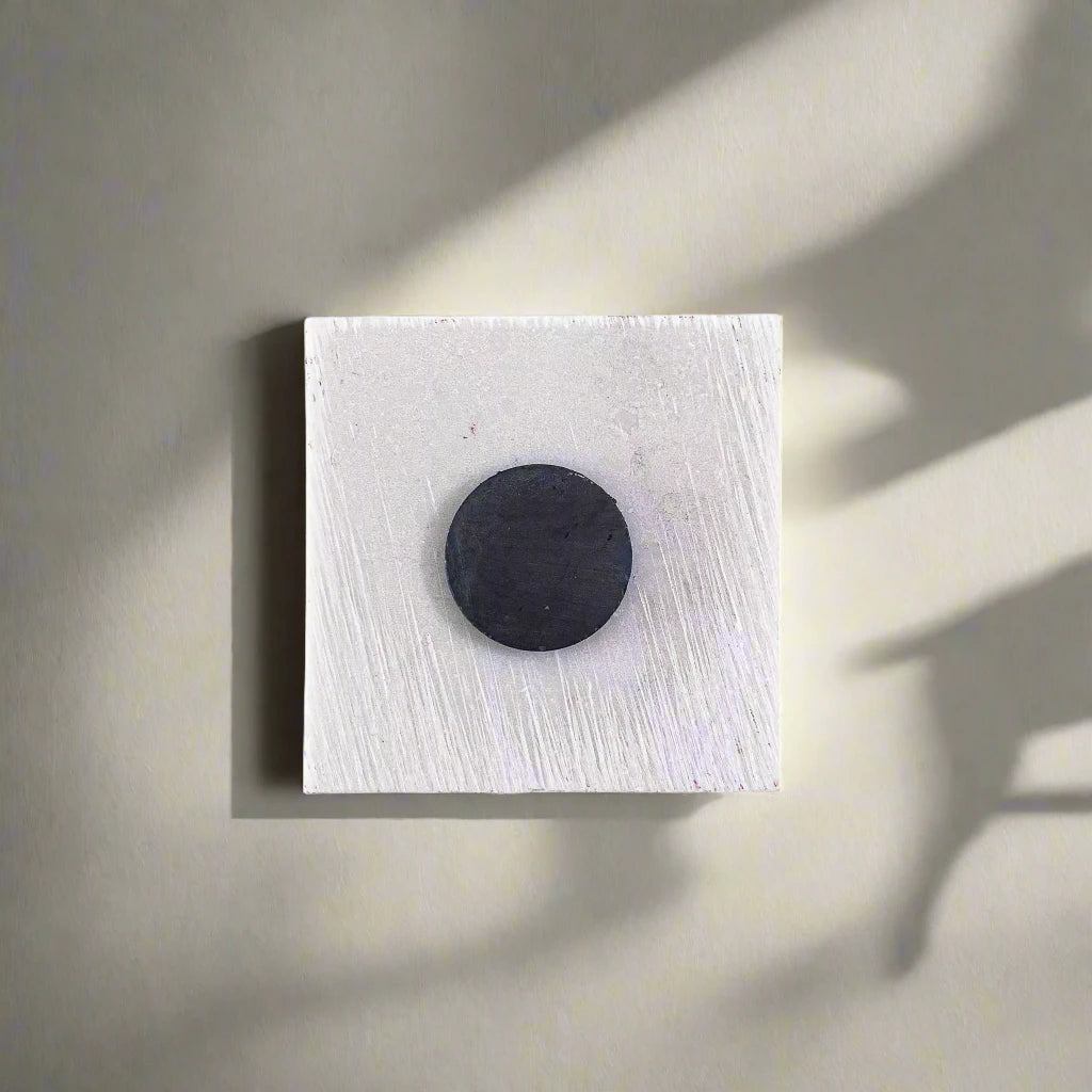 A minimalist artwork featuring a square white canvas with a small, circular, black object resembling a magnet positioned slightly above center. A faint shadow cast by the black circle extends toward the lower right corner, adding depth to the monochromatic composition.