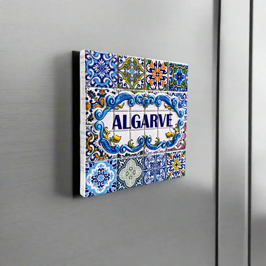 A colorful ceramic tile magnet featuring "ALGARVE" prominently in the center, surrounded by detailed blue, yellow, green, and white patterns with floral designs. Inspired by traditional Portuguese azulejos, this makes for an ideal Algarve souvenir.