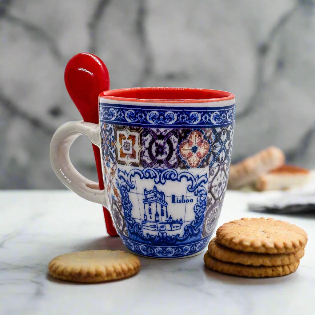 A vibrant ceramic coffee cup featuring detailed blue and red patterns with the word "Lisboa" displayed on the side. This Lisbon souvenir includes a red spoon inserted through the handle. A black, round plastic object, possibly a bottle cap, sits to the left of the mug.
