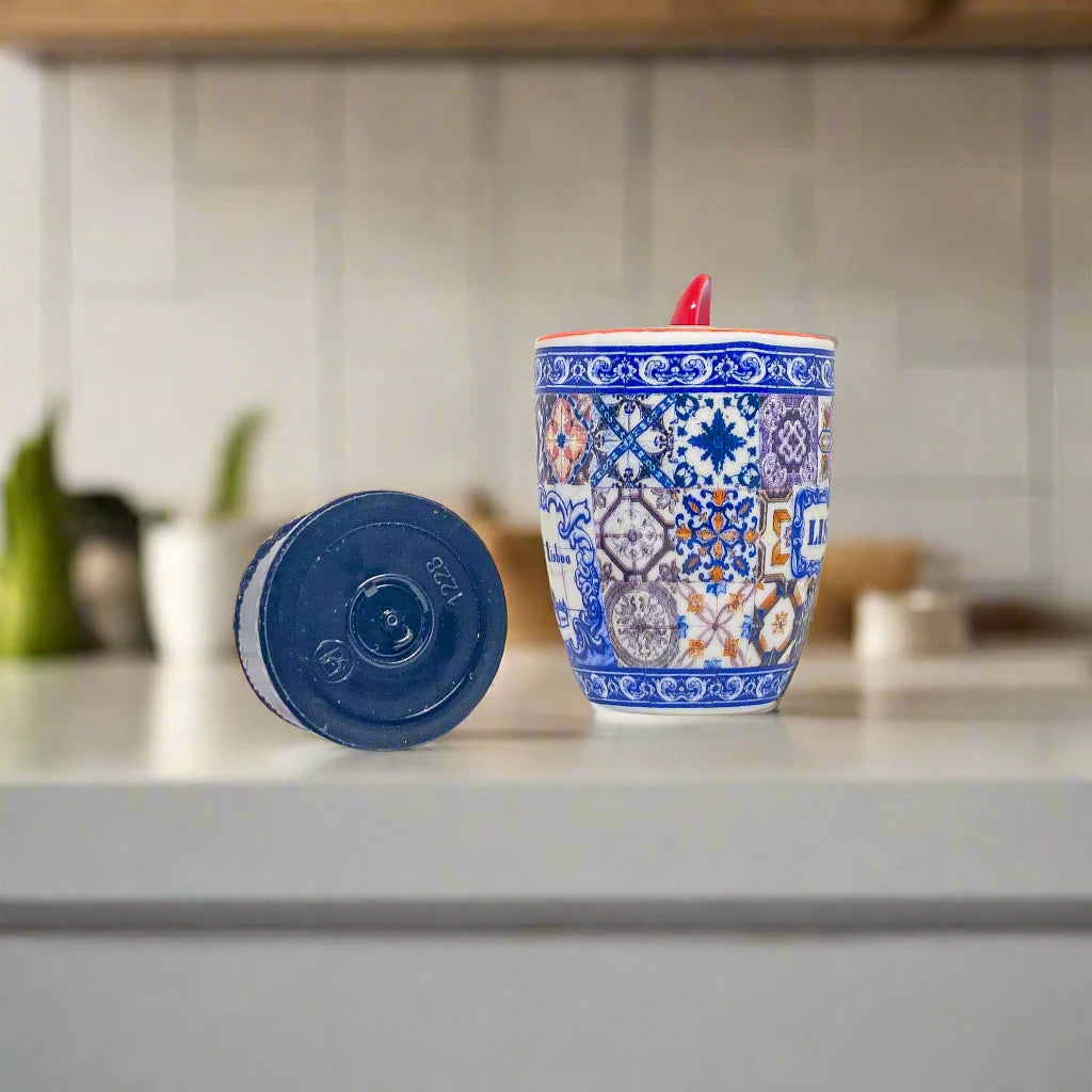 A decorative ceramic coffee mug adorned with intricate blue and red designs and labeled "Lisboa." The mug comes with a red spoon nestled in its handle, making it an ideal Lisbon keepsake. To its left, there is a small black plastic piece, likely a bottle cap, set against a white backdrop.