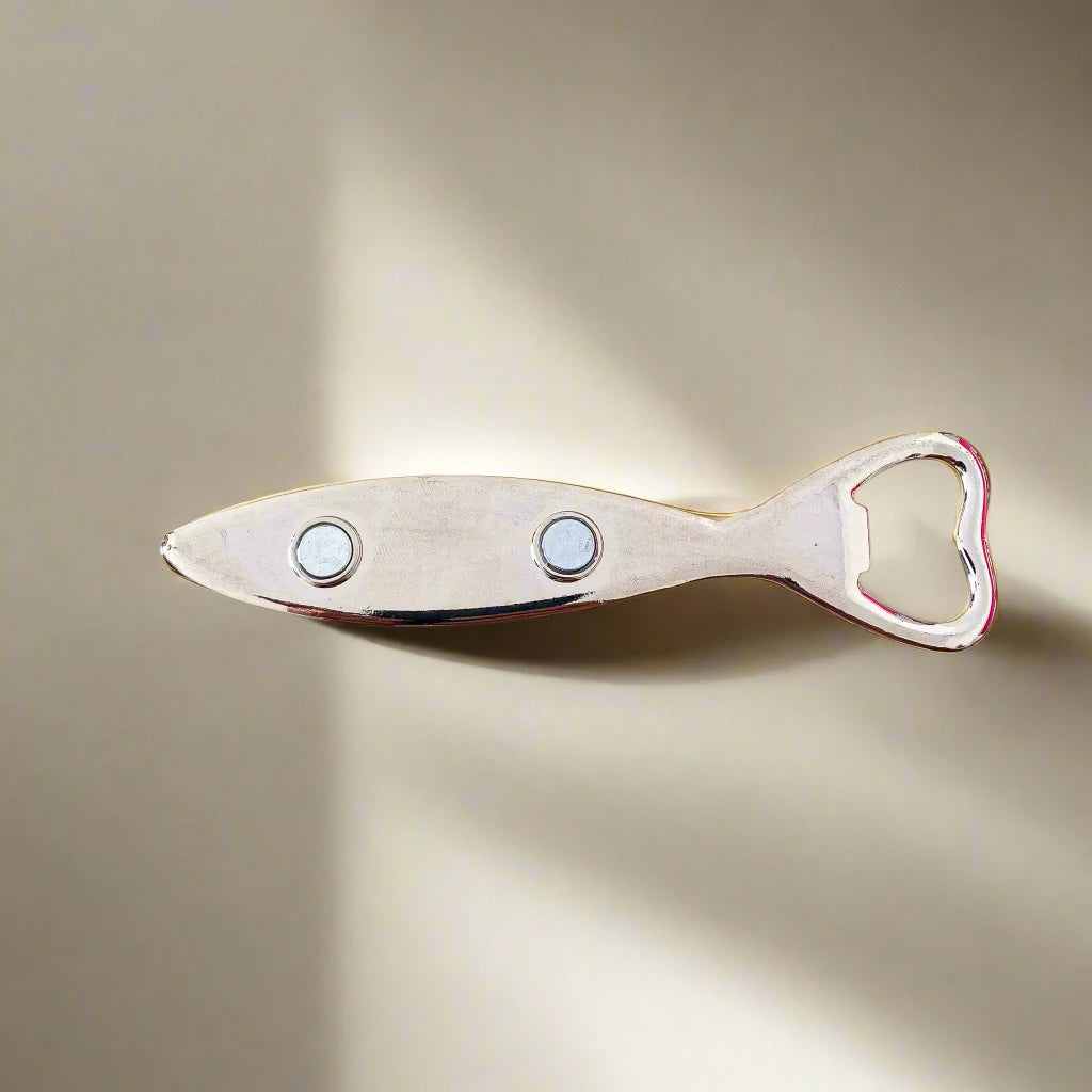 A sardine-shaped bottle opener with a detailed, colorful depiction of Lisbon, Portugal on the handle. The illustration highlights famous city landmarks, traditional Portuguese tiles, and a tram. The handle is extended with the opener located at the fish's mouth, and it also functions as a fridge magnet.