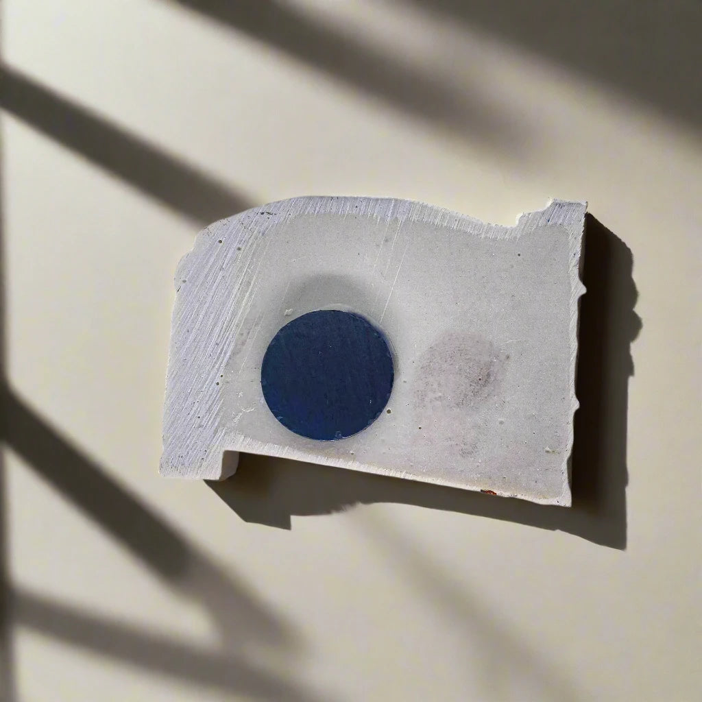 A rectangular piece of textured white material with rough edges is photographed against a neutral background. A dark blue circular shape is in the center of the material, reminiscent of a unique souvenir from Portugal. The shadow of window blinds is cast diagonally over the object.