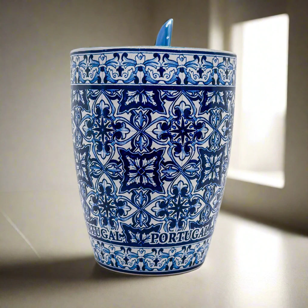 A ceramic souvenir mug decorated with intricate blue and white patterns inspired by traditional Portuguese tiles, with "PORTUGAL" elegantly encircling the base. The mug is paired with a matching blue spoon that slots conveniently into the handle.