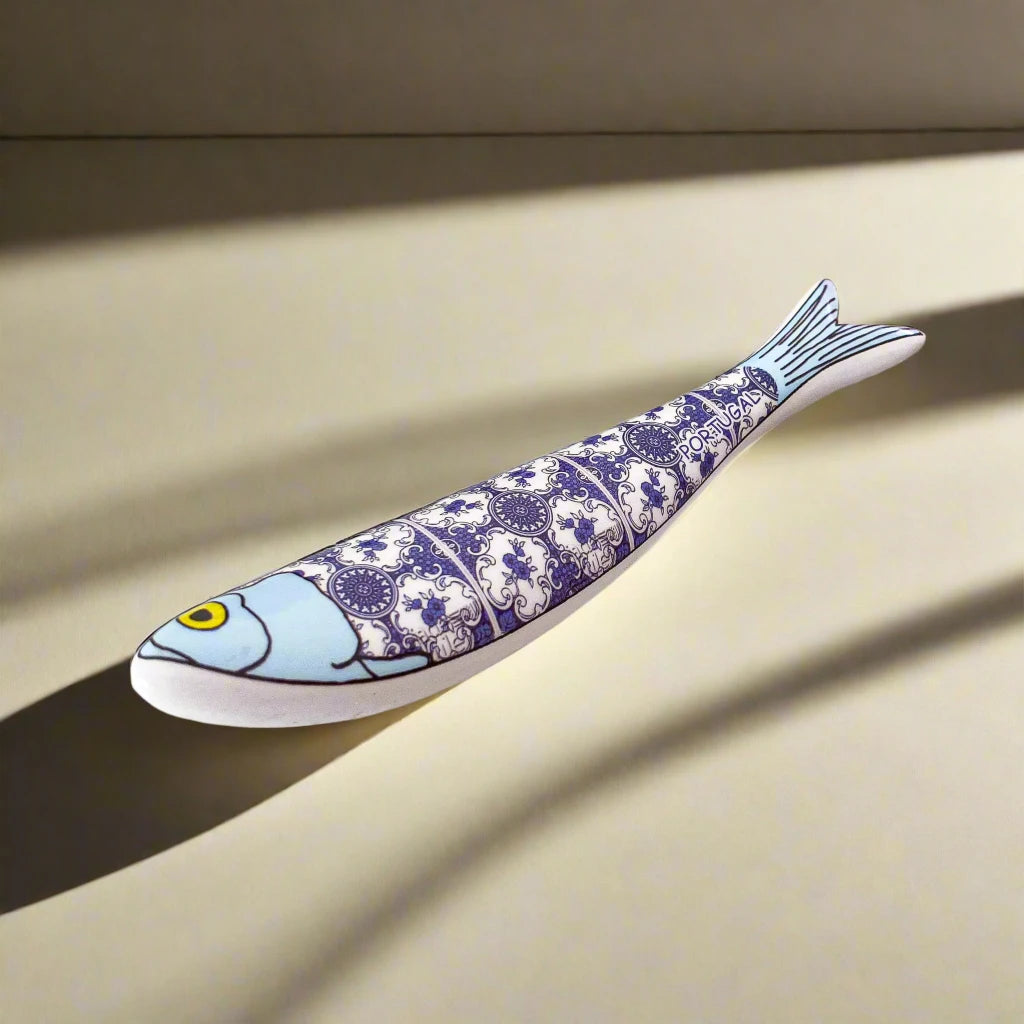 A ceramic sardine figurine adorned with a blue and white intricate pattern, inspired by traditional Portuguese tiles. The fish features a blue head with a yellow eye, and its body is segmented into four sections with the detailed pattern. The word "Portugal" is elegantly placed near the tail, making it an ideal souvenir.