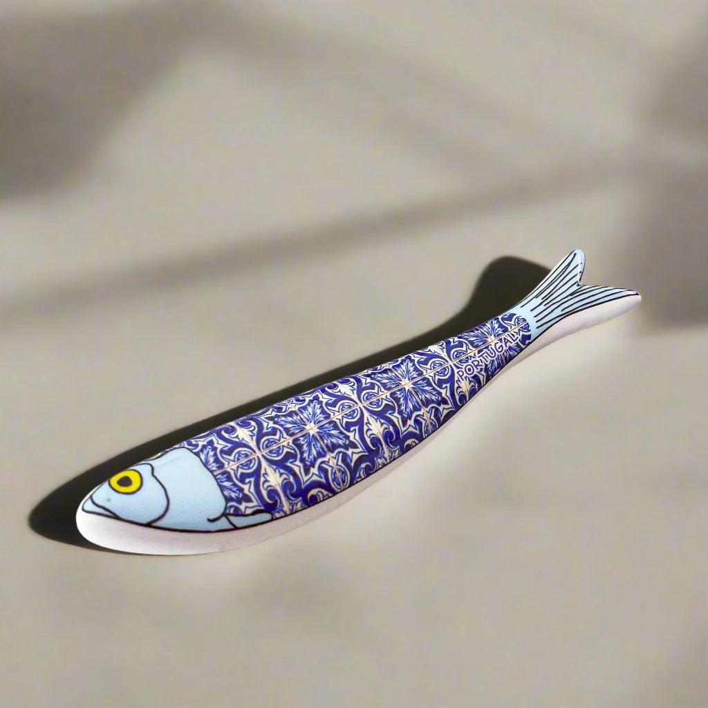 A ceramic plate shaped like a sardine, featuring a blue and white intricate pattern that echoes traditional Portuguese tile designs. The fish's face and tail are solid blue, while its body displays the decorative tile patterns. This delightful piece is an ideal Portugal-themed souvenir.