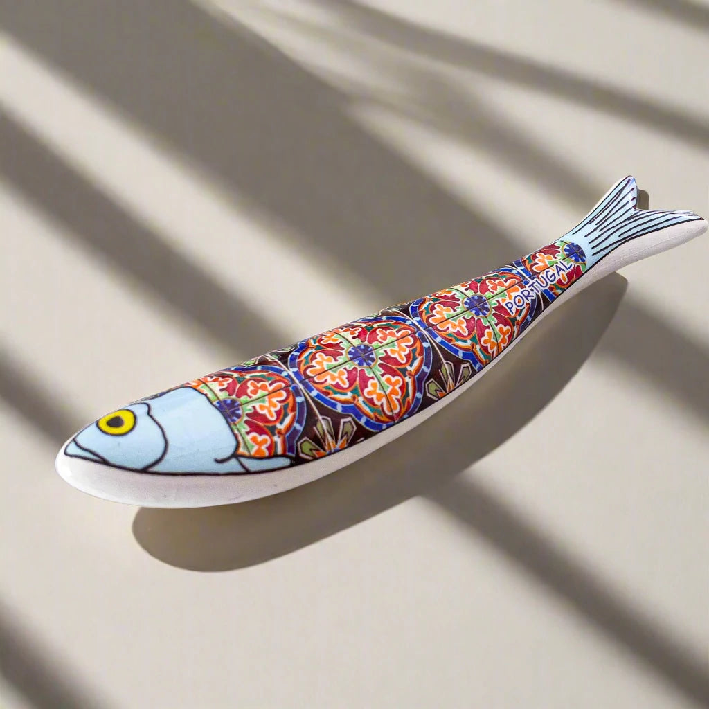 A decorative ceramic fish magnet with a blue head, yellow eye, and a detailed, colorful body pattern that resembles traditional Portuguese tiles. The body is adorned with vibrant reds, blues, and yellows, with the word "Portugal" displayed near the tail.