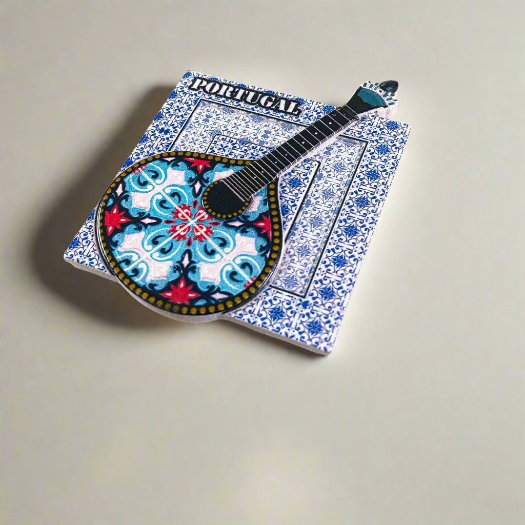 A magnet displaying the word "Portugal" at the top, with a traditional Portuguese "guitarra classica" as the central feature. The guitar is decorated with vibrant blue, red, and black patterns, highlighting its ornate design. The backdrop is a white square embellished with intricate blue tile patterns, evoking the charm of Portuguese azulejos.