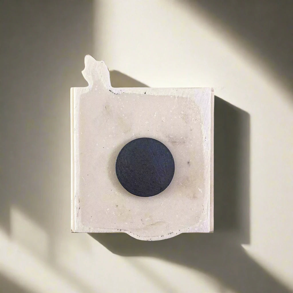 A minimalist sculpture reminiscent of a cherished souvenir: a flat, unevenly shaped white marble slab with rough edges. At its center is a perfectly round, dark circular disk. The piece, evoking memories of Portugal's artistry, casts subtle shadows on a light gray background.