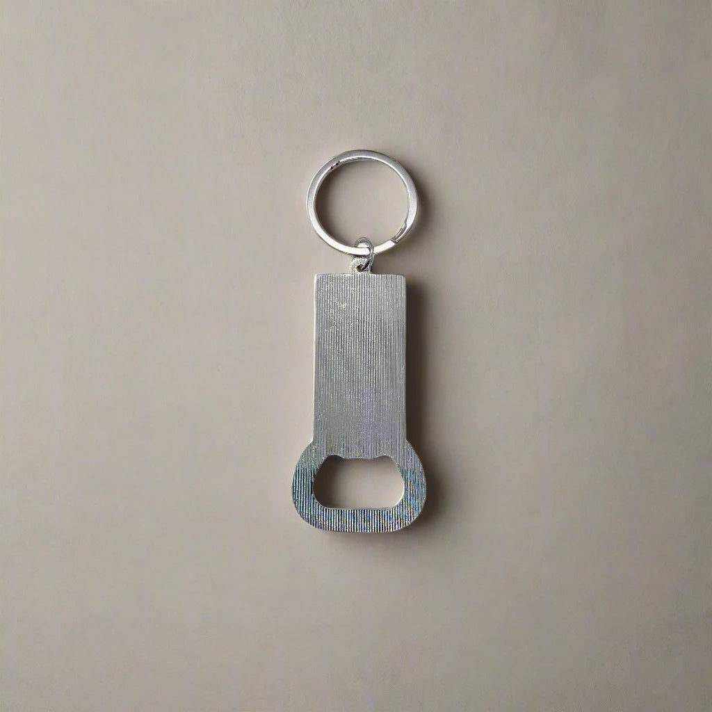 A silver keychain with a built-in bottle opener, lying on a beige surface. The keychain features a rectangular body with a circular hole at the top, attached to a key ring, and a bottle opener at the bottom—an ideal Portugal souvenir.