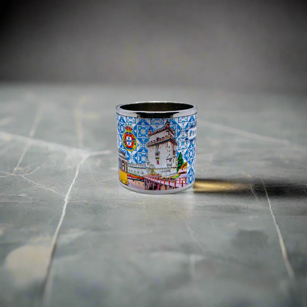 A small metal mug showcases the word "Portugal" against a blue and white tile-inspired pattern, reminiscent of traditional Portuguese azulejos. The design is accented by vibrant illustrations, including the iconic Portuguese rooster, making it a charming addition alongside your favorite Portugal-themed magnets.
