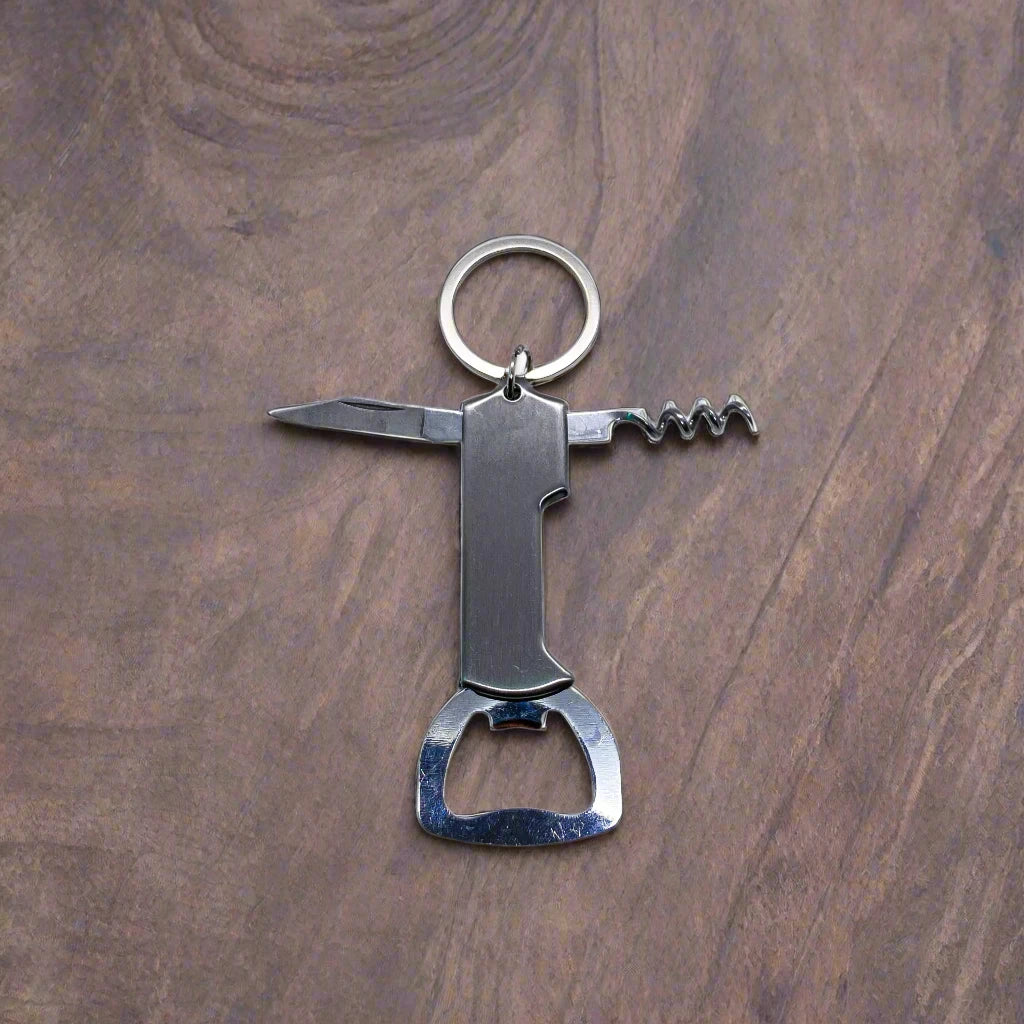 A multi-functional keychain against a wooden background. This souvenir from Portugal features a small knife blade, a corkscrew, and a bottle opener, all made of metal. The keyring is attached to the top of the tool.