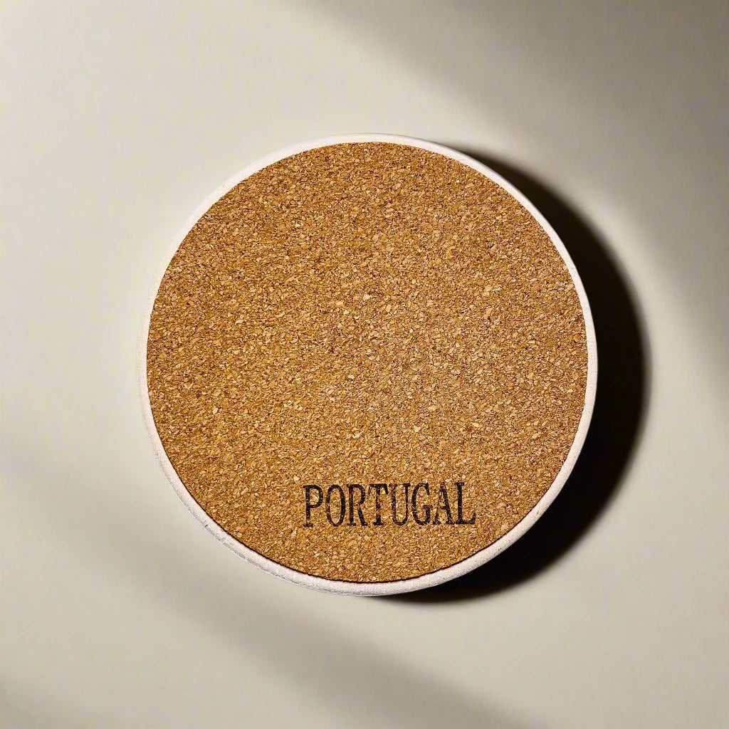 A round cork coaster with the word "PORTUGAL" inscribed on it sits on a light-colored surface. The coaster has a natural, textured appearance, typical of cork material. This charming souvenir is well-lit, highlighting the texture of the cork.