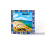 A colorful, blue-bordered ceramic tile featuring an aerial view of a busy beach in Algarve, Portugal. The beach is filled with people and umbrellas, set against a vivid blue sky and turquoise sea. The word "ALGARVE" is prominently displayed at the top.