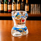 Angled front view of Albufeira women bikini style shot glass featuring Algarve beach artwork, palm trees, sailboats, and I Love Albufeira print on wooden bar counter.
