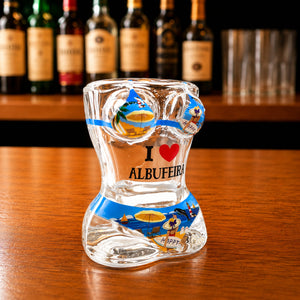 Angled front view of Albufeira women bikini style shot glass featuring Algarve beach artwork, palm trees, sailboats, and I Love Albufeira print on wooden bar counter.