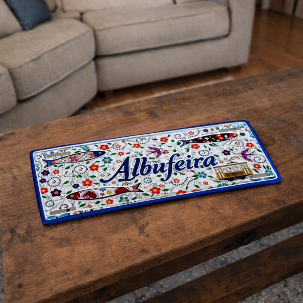 Albufeira metal souvenir sign placed on a wooden table showing floral motifs, stylized sardines and a classic tram illustration
