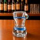Side profile of transparent Albufeira souvenir shot glass featuring blue decorative stripe and Algarve beach-inspired bikini artwork on wooden surface.