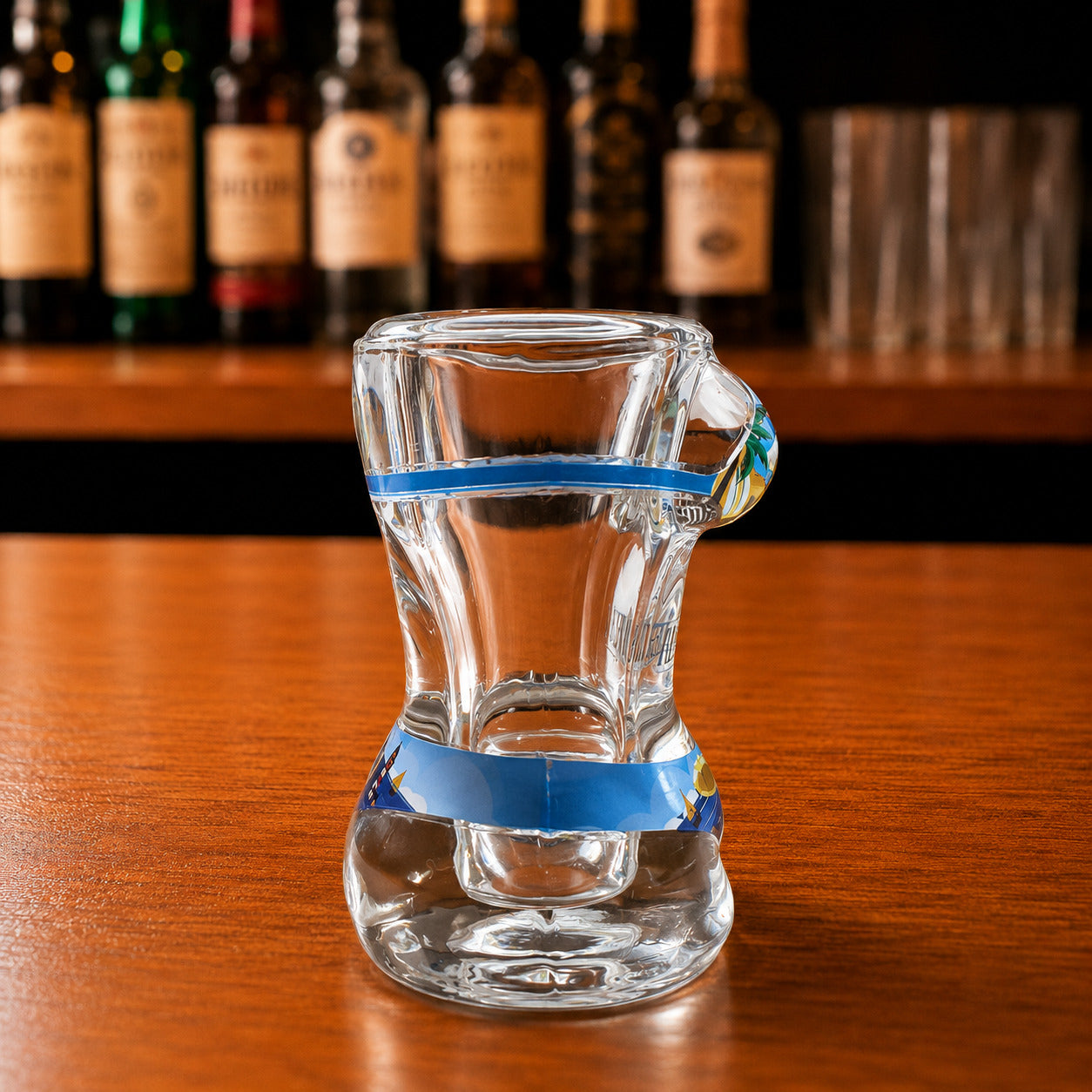 Side profile of transparent Albufeira souvenir shot glass featuring blue decorative stripe and Algarve beach-inspired bikini artwork on wooden surface.