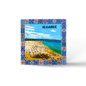 A colorful, blue-bordered ceramic tile featuring an aerial view of a busy beach in Algarve, Portugal. The beach is filled with people and umbrellas, set against a vivid blue sky and turquoise sea. The word "ALGARVE" is prominently displayed at the top.