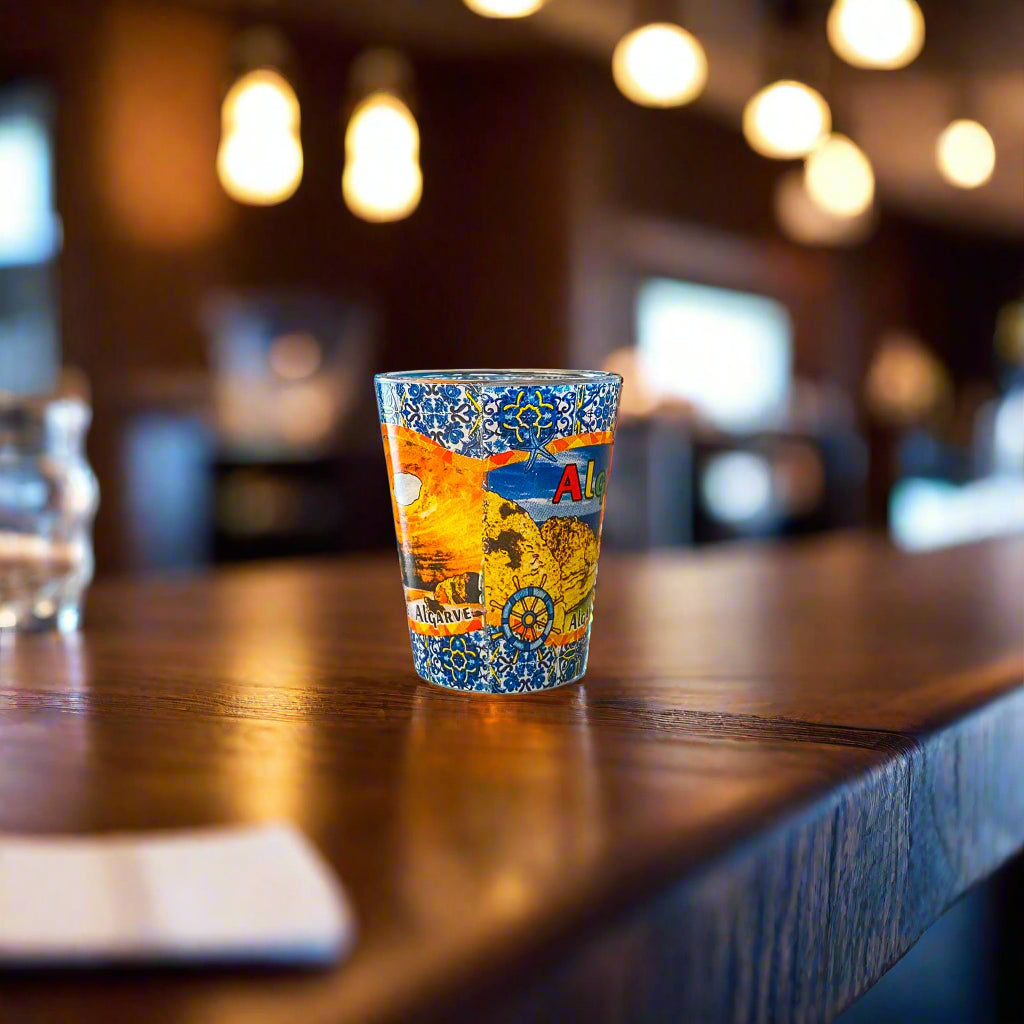 Algarve Portugal souvenir shot glass decorated with famous coastal cliffs, ocean scenery, and blue azulejo tile designs displayed on a wooden bar counter.