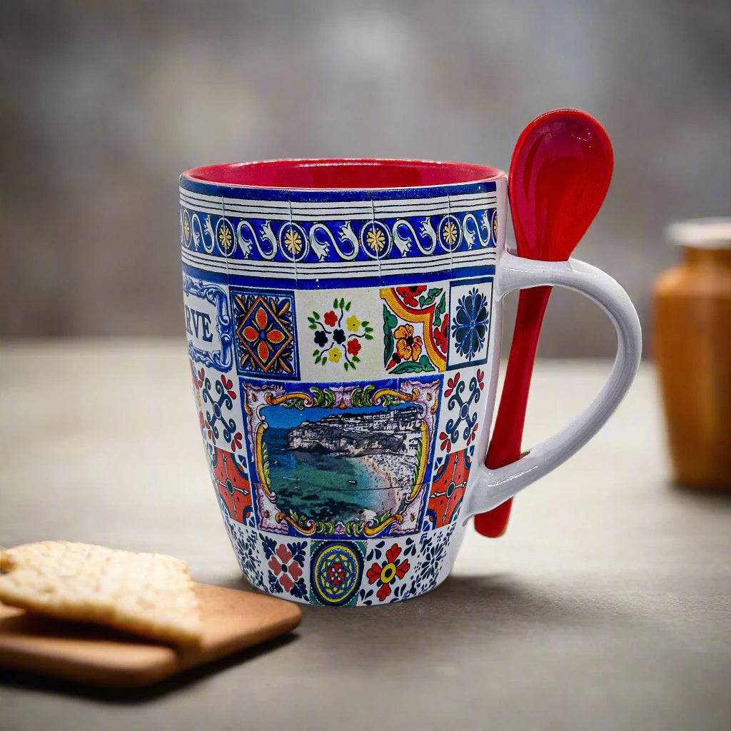 A colorful ceramic mug with intricate patterns and a scenic landscape image of the Algarve on the front sits on a table. The souvenir mug has a red interior and comes with a red spoon tucked into the handle. Two square crackers are placed on a wooden board nearby.