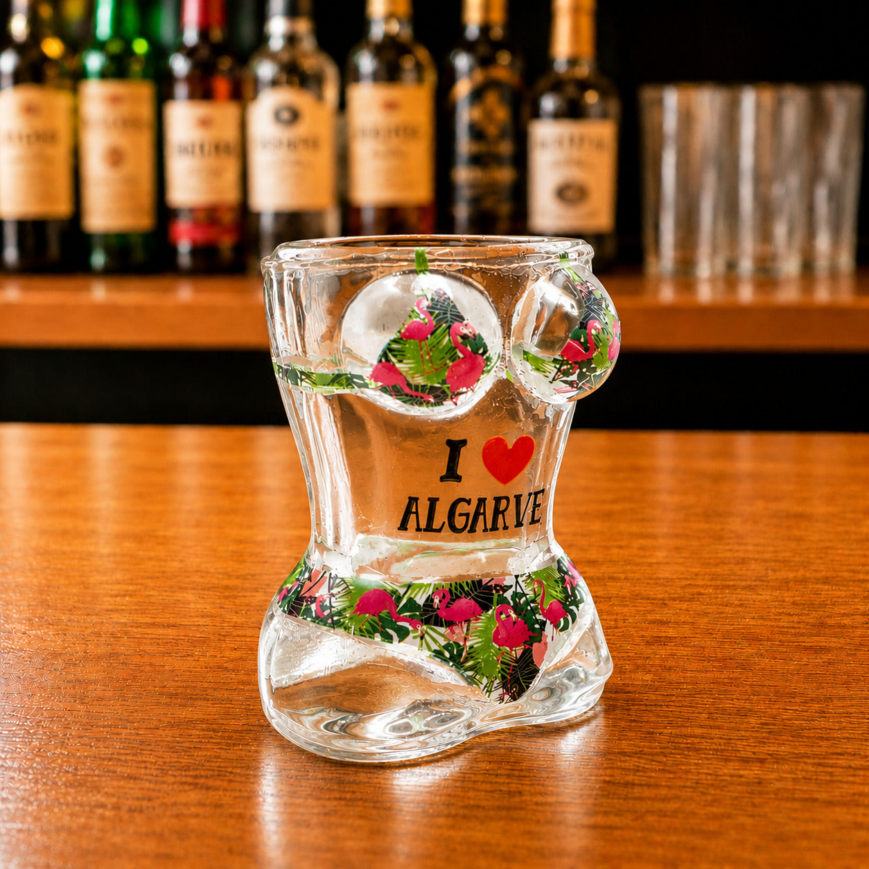 Angled front view of Algarve flamingo tropical bikini style shot glass featuring pink flamingo artwork, tropical foliage patterns, and I Love Algarve text on wooden surface.