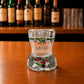 Back view of Algarve flamingo tropical bikini style shot glass with tropical leaf patterns, pink flamingo illustrations, and green decorative stripe on wooden bar counter.