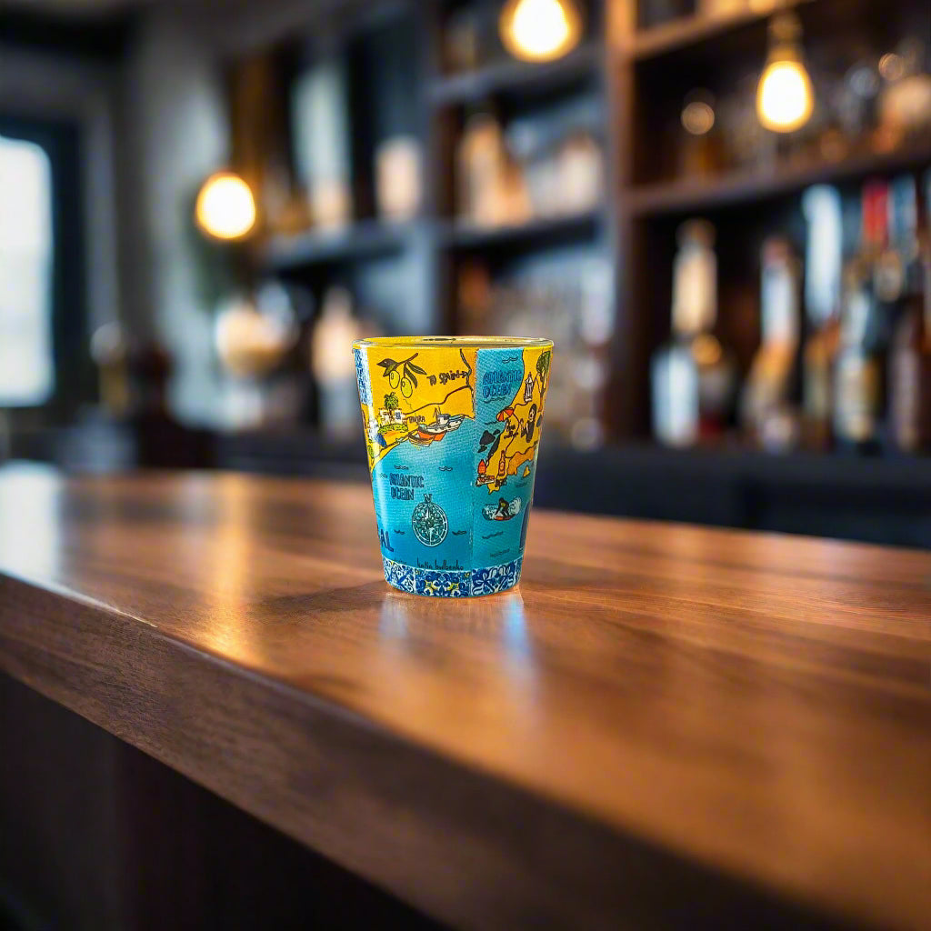 Colorful Algarve souvenir shot glass featuring illustrated Portugal coastal map artwork in a warm bar setting.