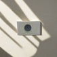 A small white rectangular block, resembling a souvenir magnet, with a dark circular hole in the center is mounted on a light-colored wall. Shadows from nearby objects fall across the wall and the block, creating diagonal patterns of light and shade.