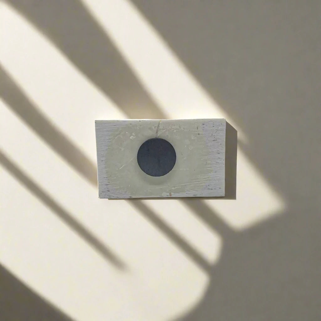 A small white rectangular block, resembling a souvenir magnet, with a dark circular hole in the center is mounted on a light-colored wall. Shadows from nearby objects fall across the wall and the block, creating diagonal patterns of light and shade.