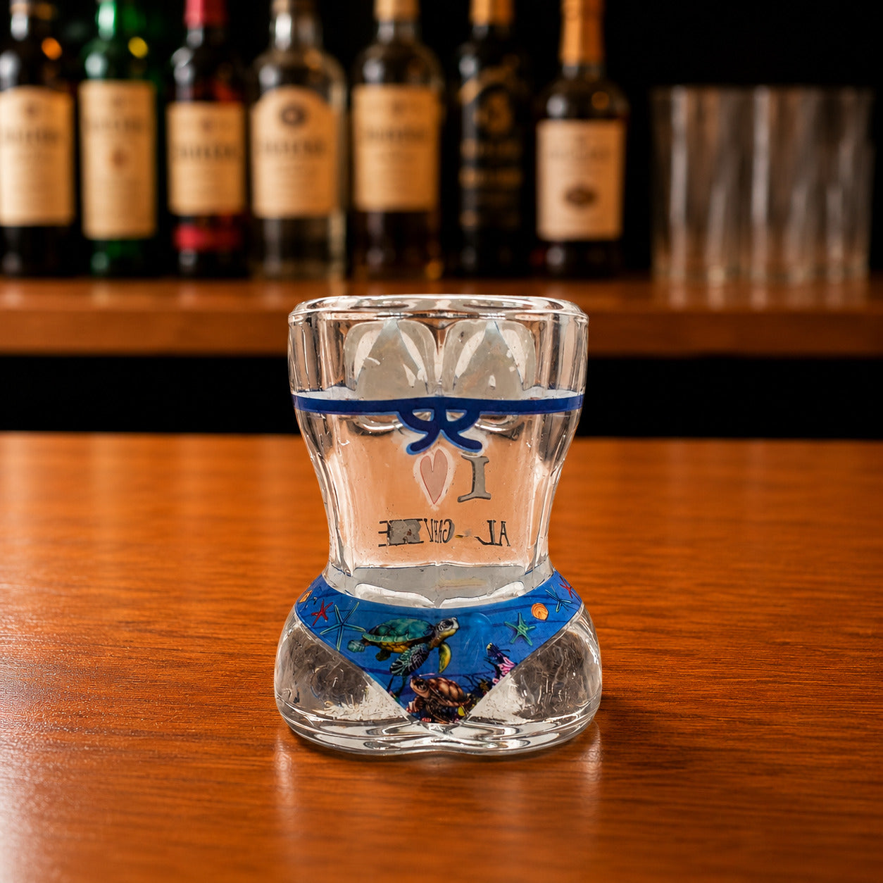 Back view of Algarve sea turtle ocean bikini style shot glass with blue underwater artwork, sea turtle illustrations, and decorative marine design on wooden bar counter.