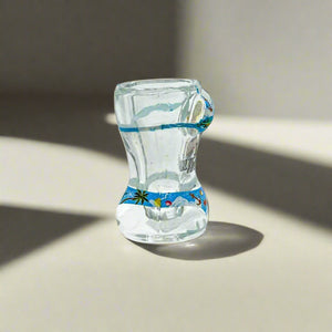 A clear shot glass designed with a sculpted torso in a bikini, showcasing beach and ocean imagery. The bikini is illustrated with scenes of the seaside. The front of the glass displays "I ♥ Algarve" in bold black and red letters, making it an ideal keepsake from the Algarve region.