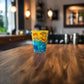 Portugal Algarve shot glass decorated with illustrated coastal landmarks and regional icons photographed on a rustic wooden table.