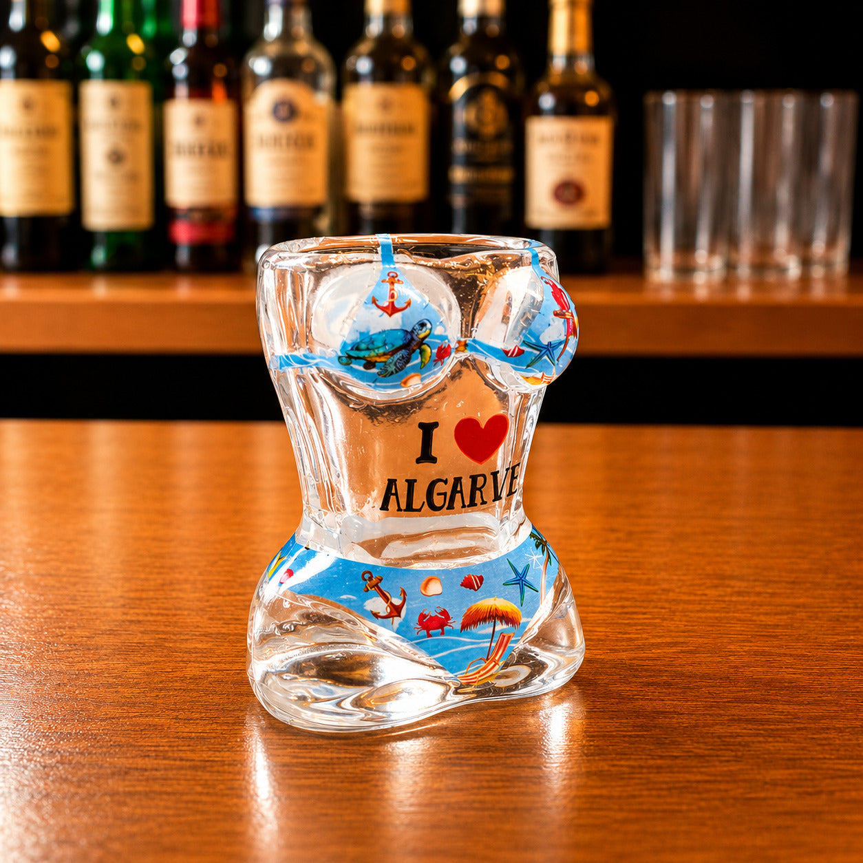 Angled front view of Algarve tropical beach bikini style shot glass with sea turtle artwork, tropical beach illustrations, and I Love Algarve text on wooden bar counter.