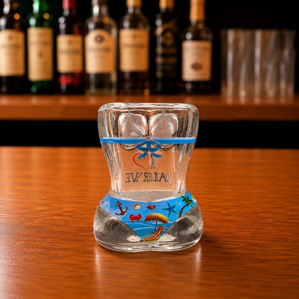 Back view of Algarve tropical beach bikini style shot glass featuring seaside illustrations with beach chair, palm tree, starfish, anchor, and summer ocean artwork on wooden bar counter.