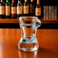 Side profile of transparent Algarve souvenir shot glass with tropical beach-themed bikini artwork, blue decorative stripe, and seaside illustrations on wooden surface.