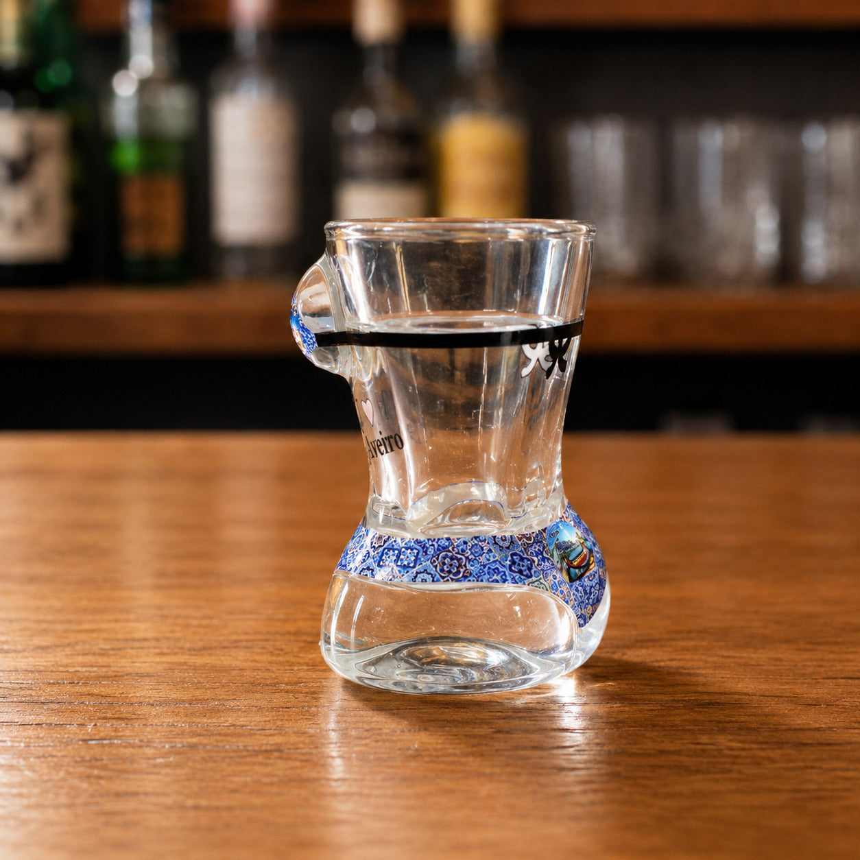 Side profile of transparent Aveiro souvenir shot glass featuring blue azulejo bikini artwork, black stripe detail, and Portuguese coastal imagery.