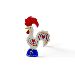 Image of a colorful ceramic rooster figurine known as the Rooster of Barcelos, a cultural symbol and popular souvenir from Portugal. The rooster has red details on its comb, wattle, and heart-shaped accents on its white body, with "Portugal" written on the blue base. Ideal for use as a decorative magnet.
