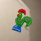 A vibrant decorative rooster figurine from Portugal, showcasing a green body with red hearts and white dots, along with a red comb and wattles. It stands on a blue base with "Portugal" inscribed, and doubles as a magnet, making it a perfect souvenir.