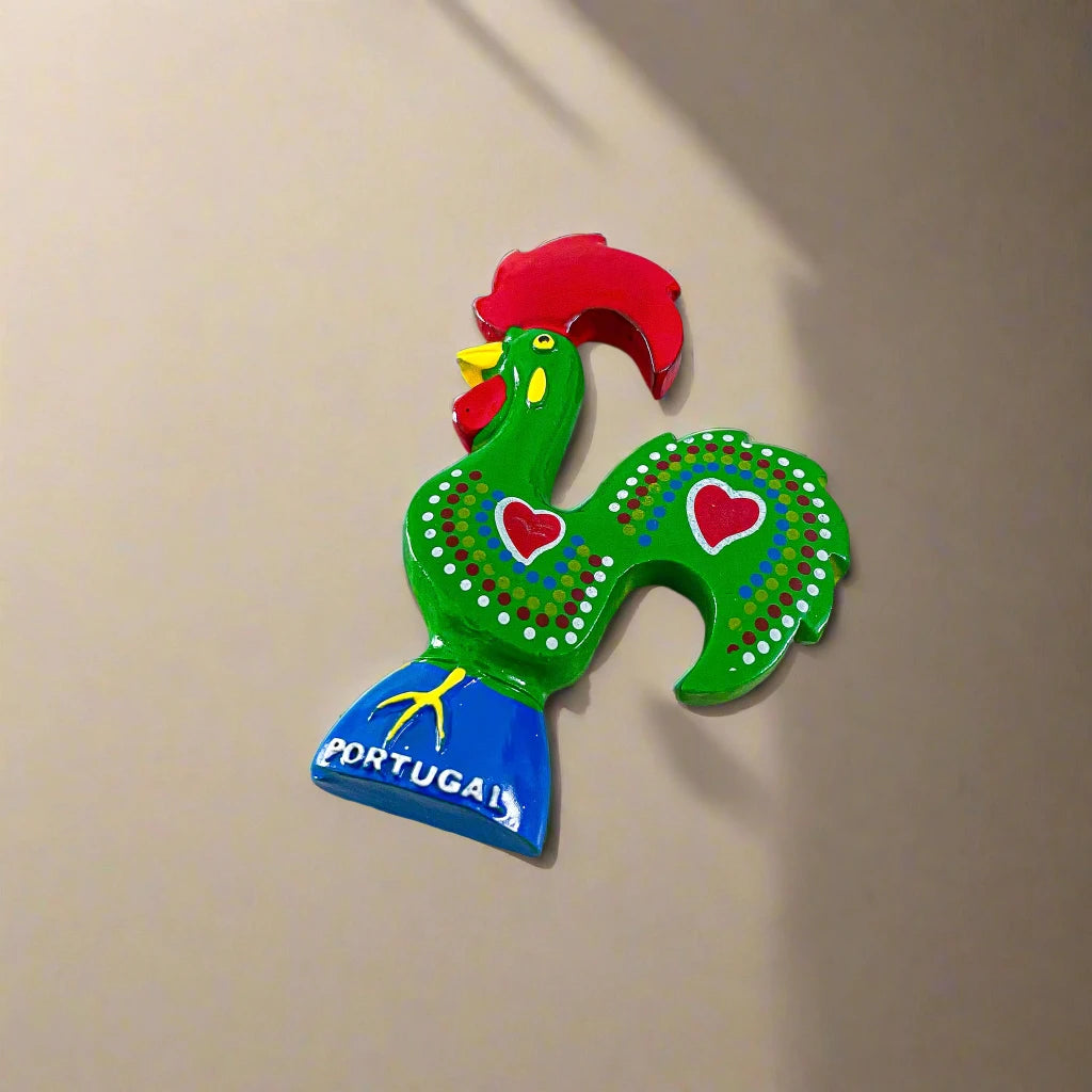 A vibrant decorative rooster figurine from Portugal, showcasing a green body with red hearts and white dots, along with a red comb and wattles. It stands on a blue base with "Portugal" inscribed, and doubles as a magnet, making it a perfect souvenir.
