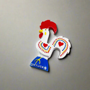 Colorful ceramic rooster figurine, the Rooster of Barcelos, a beloved symbol of Portuguese culture. It features red accents on its comb, wattle, and heart-shaped patterns on a white body, with "Portugal" on the blue base. Perfect as a decorative magnet.